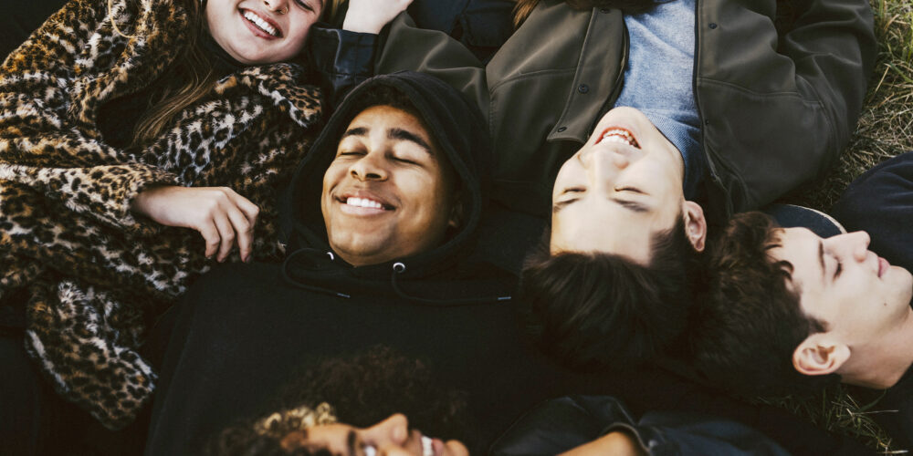 Ett gäng unga personer ligger på marken med sina huvuden ihop och skrattar och ler.
