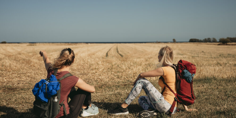 Två personer som sitter vid ett fält och pekar mot horisonten. Båda bär träningskläder och ryggsäckar.
