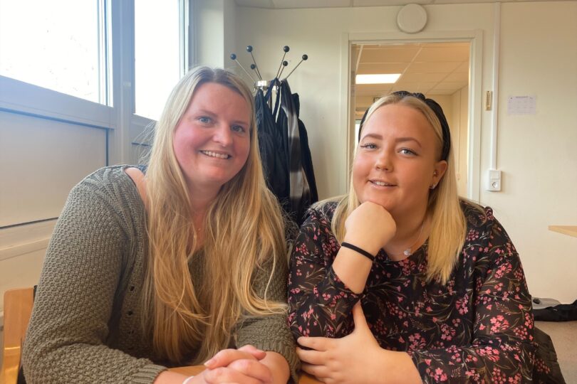 Två personer som ler. Båda två har blont hår. De sitter ner runt ett bord.