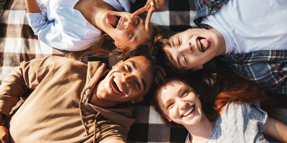 Unga människor av olika etniciteter, både killar och tjejer, skrattar och ligger på en filt i en cirkel utomhus.