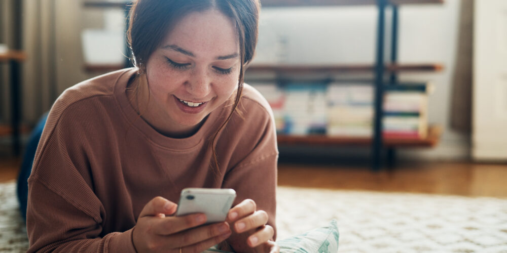 Tonåring ligger på mage på golvet och tittar på sin telefon.