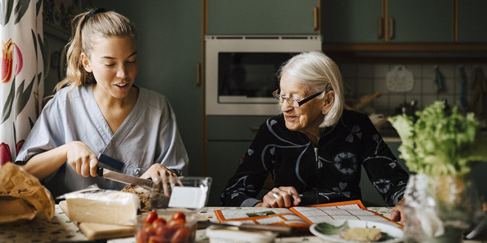 En äldre kvinna sitter tillsammans med en hemtjänstpersonal vid köksbordet och förbereder frukost.