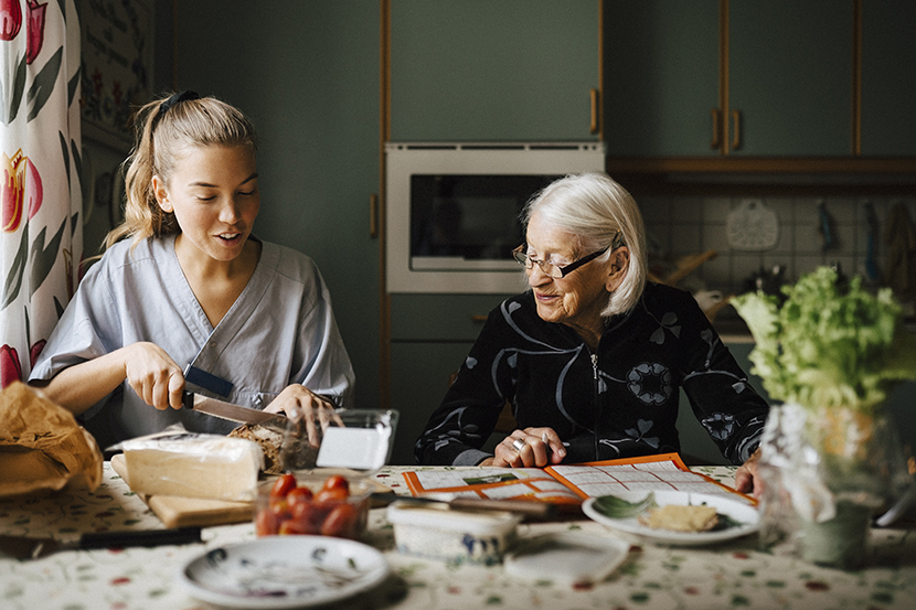 En äldre kvinna sitter tillsammans med en hemtjänstpersonal vid köksbordet och förbereder frukost.