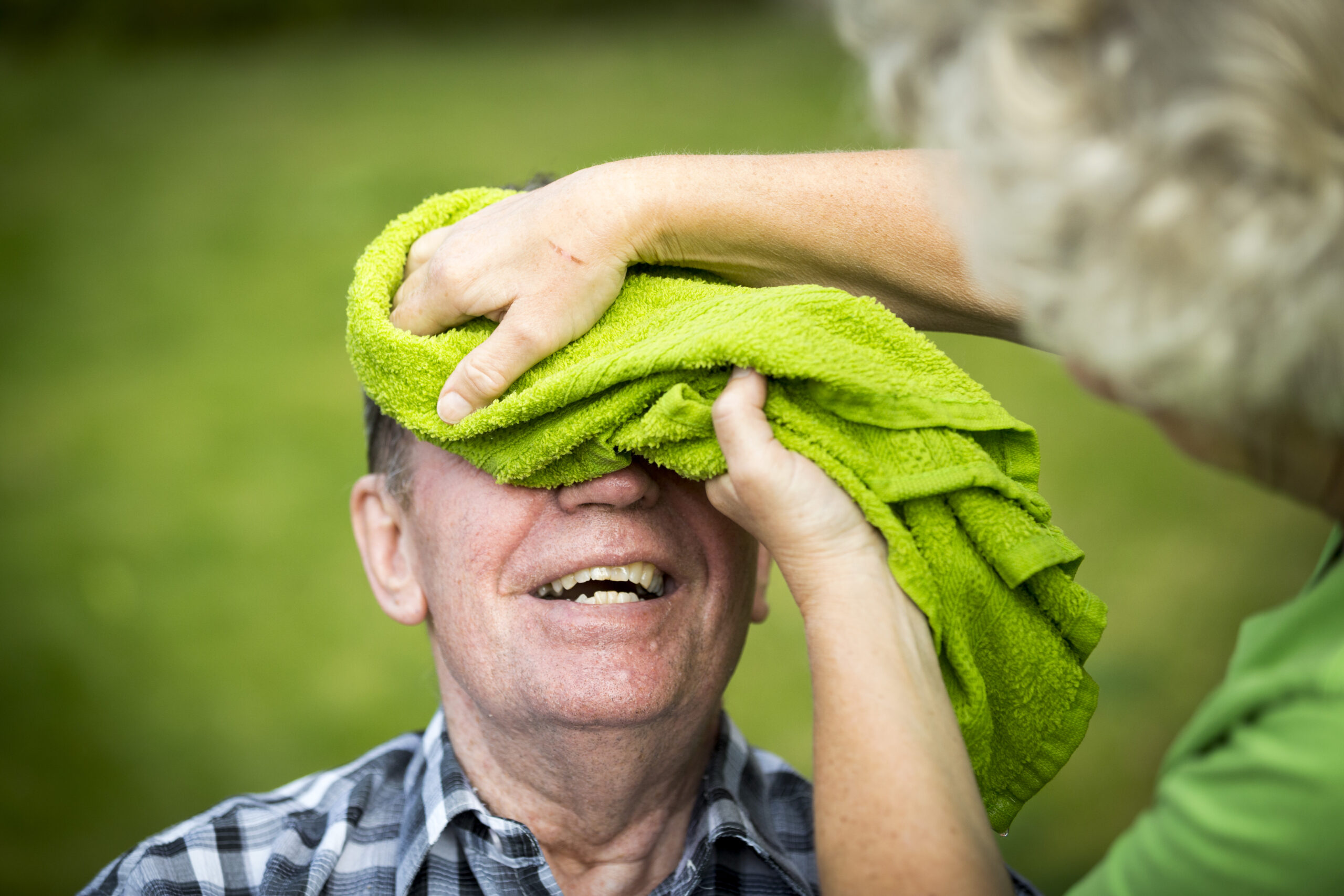 En äldre ljushyad kvinna baddar pannan på en ljushyad lätt rosig äldre man med grön våt handduk utomhus.
