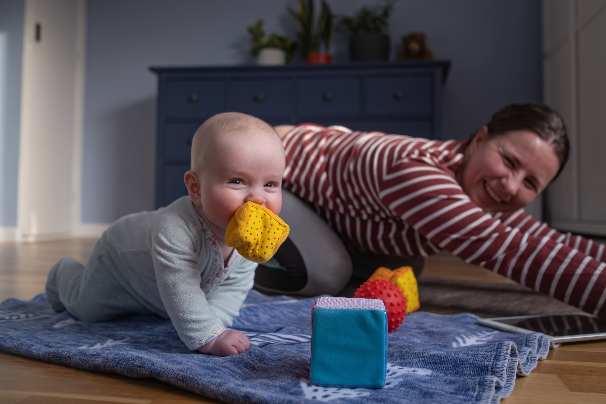 En kvinna gör en yogaövning på golvet tillsammans med ett litet barn. Bebisen som har en mjuk leksak i munnen tittar in i kameran med busig blick.