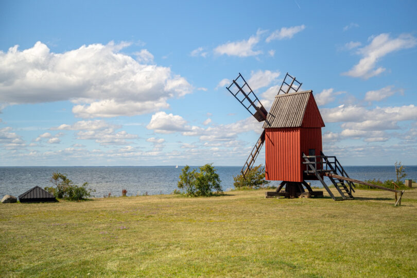 En röd kvarn på en grön gräsmatta riktad mot de blåa havet en solig dag med små lätta moln. Foto: UpZoneStudios