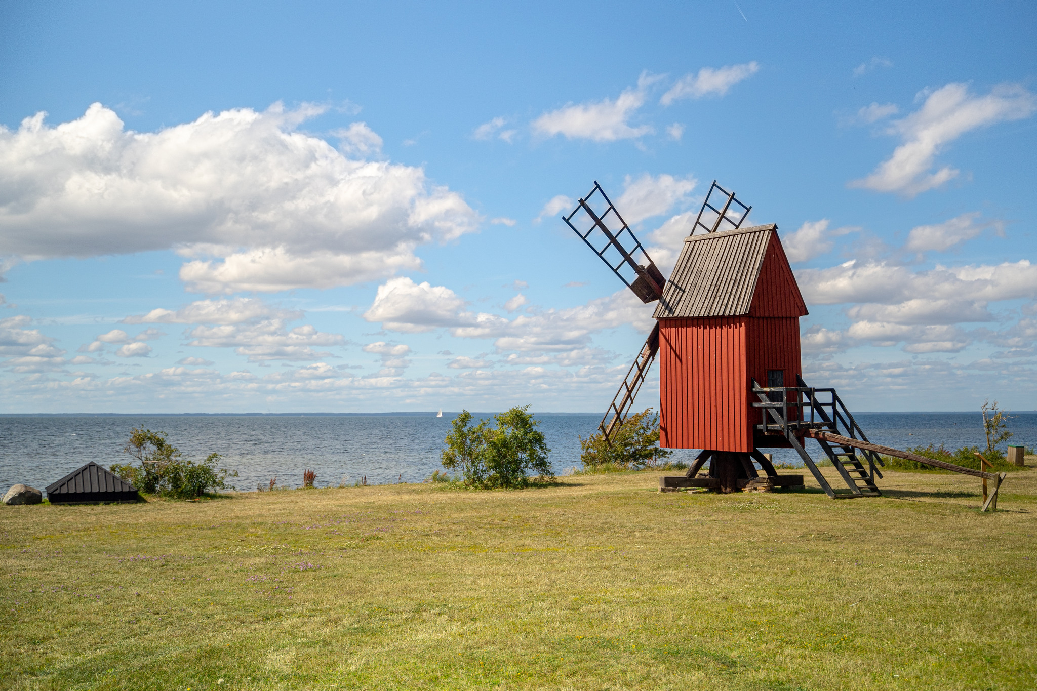 En röd kvarn på en grön gräsmatta riktad mot de blåa havet en solig dag med små lätta moln. Foto: UpZoneStudios