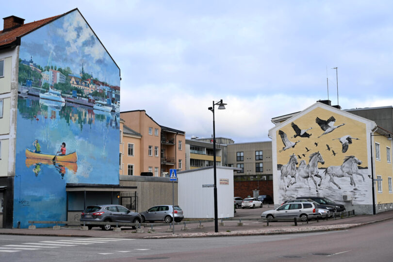 Väggmålningar på två byggnader i stadsmiljö föreställande ett par som paddlar kanot i en blå stadsmiljö samt galopperande hästar och tranor mot en gul bakgrund.