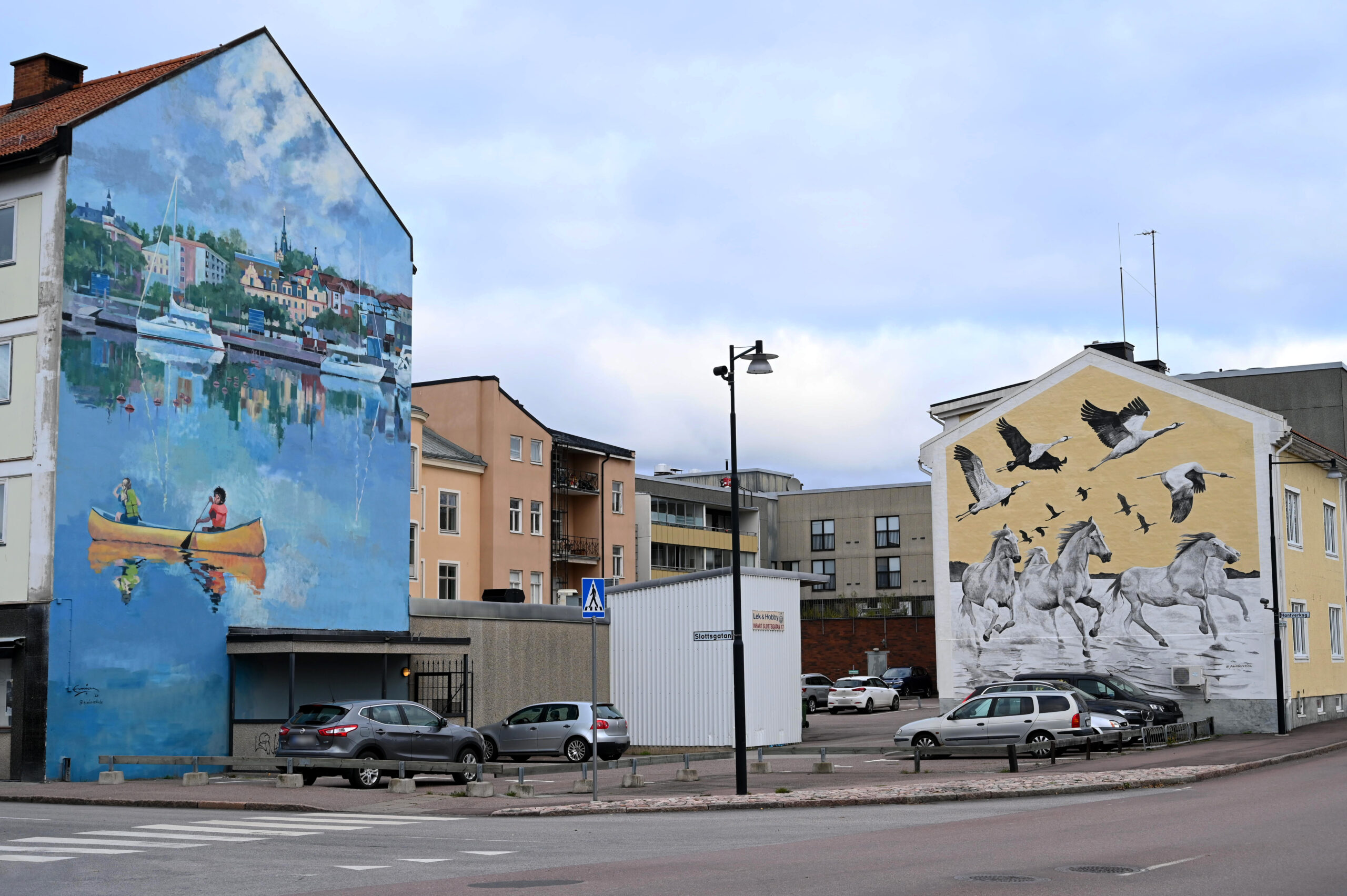 Väggmålningar på två byggnader i stadsmiljö föreställande ett par som paddlar kanot i en blå stadsmiljö samt galopperande hästar och tranor mot en gul bakgrund.