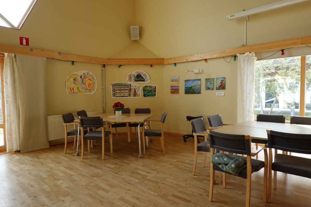 Gemensamhetslokal med konst på väggarna bakom bord och stolar.