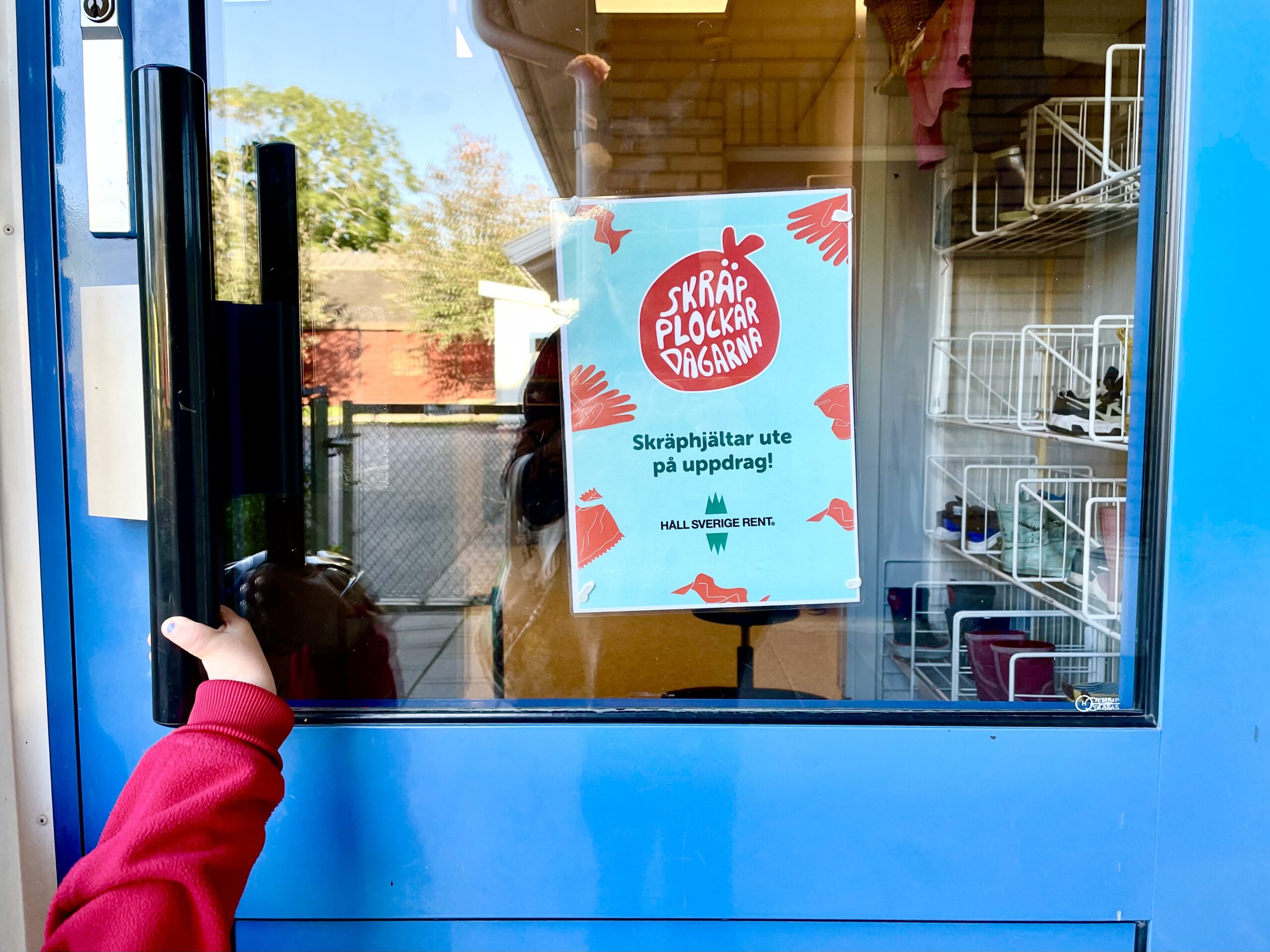 Blå dörr till förskola med en affisch på framsidan. En liten barnhand håller i handtaget.