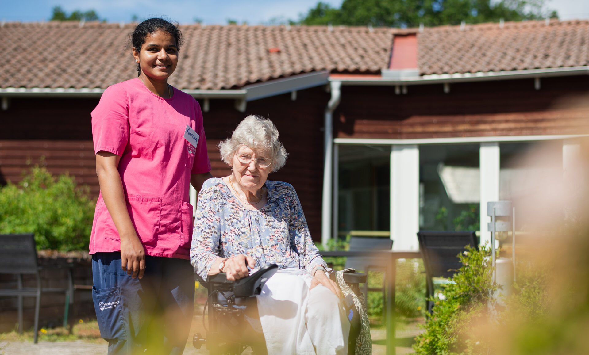 En äldre gråhårig dam sitter i en rullstol och ler mot kameran. Till vänster om henne står en leende mörkhyad ung kvinna i rosa och mörkblå vårdkläder med ena handen på rollstolens rygg. Solen skiner och i bakgrunden syns gröna buskar och svarta utemöbler framför en röd träbyggnad med stora fönster och tegeltak.