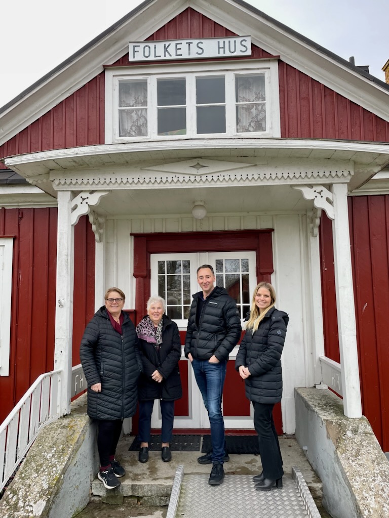 Fyra personer, tre kvinnor och en man, står i vinterkläder på farstun till ett rött hus med vita knutar och ler mot kamera.
