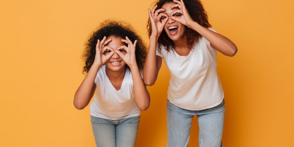 Porträtt av två mörkhyade systrar med vita t-shirts som skrattar mot en studiobakgrund i orange.