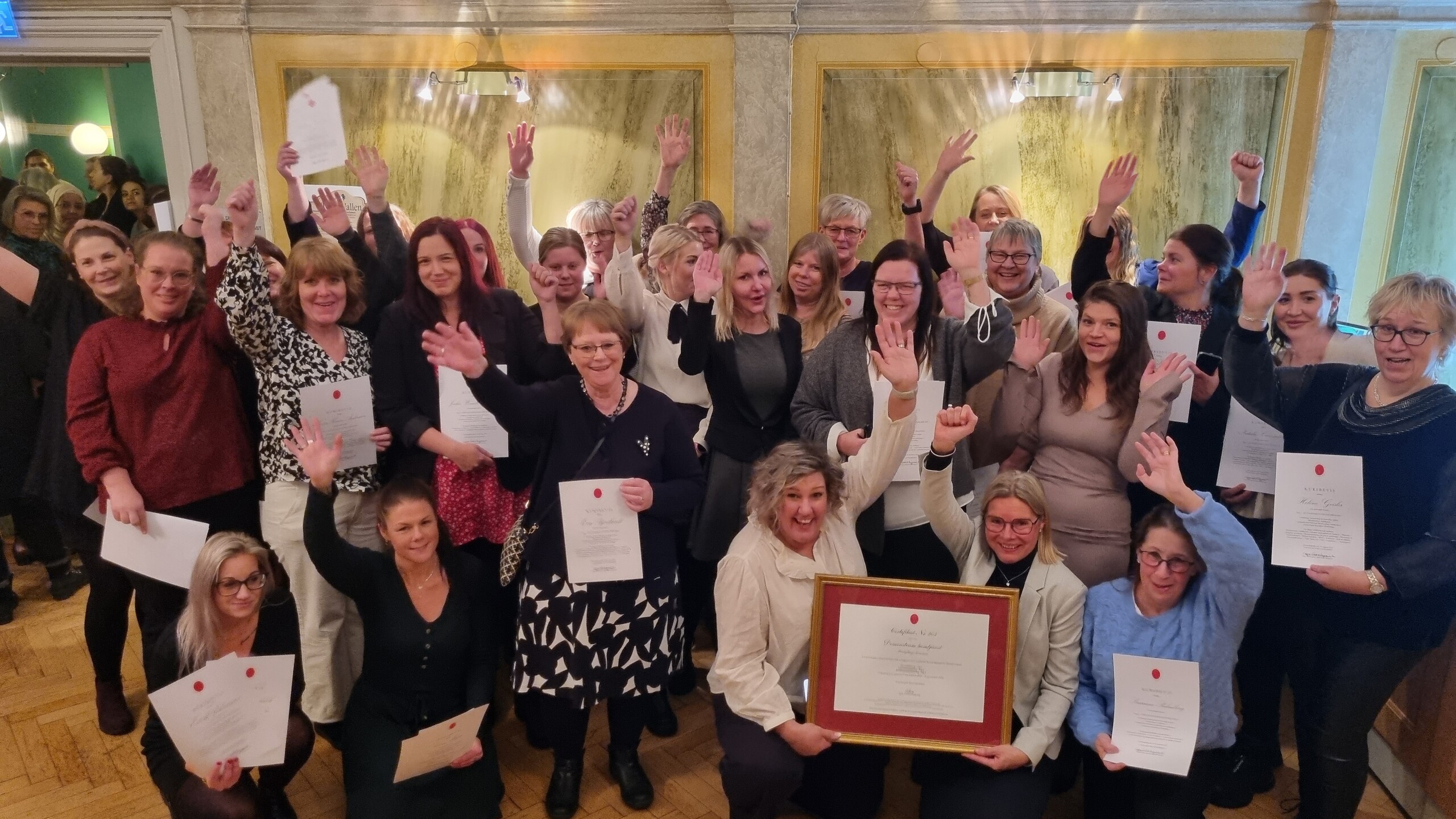 En grupp om ca 30 personer håller upp certifikat och diplom och ler mot kameran.