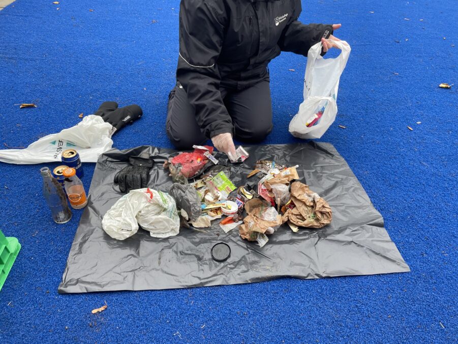 En person, på vilken man inte ser huvudet, sitter på knä på ett blått golv. Framför ligger en presenning utbredd där olika sorters skräp radats upp.