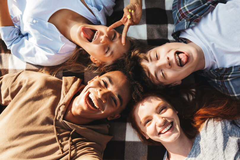 Unga människor av olika etniciteter, både killar och tjejer, skrattar och ligger på en filt i en cirkel utomhus.