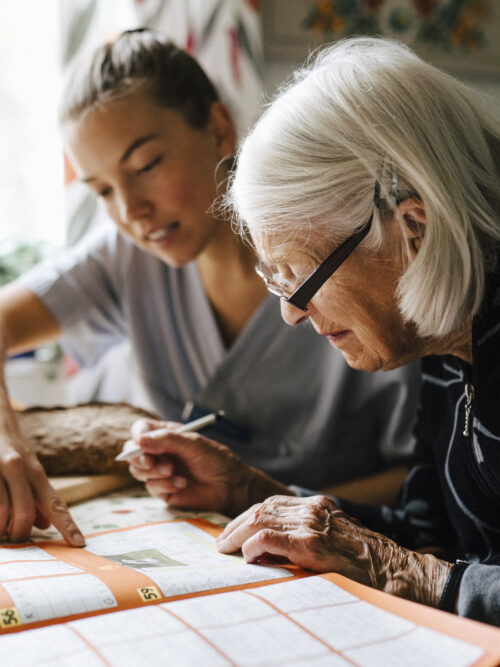En äldre kvinna siter och löser korsord med en kvinna från hemtjänsten