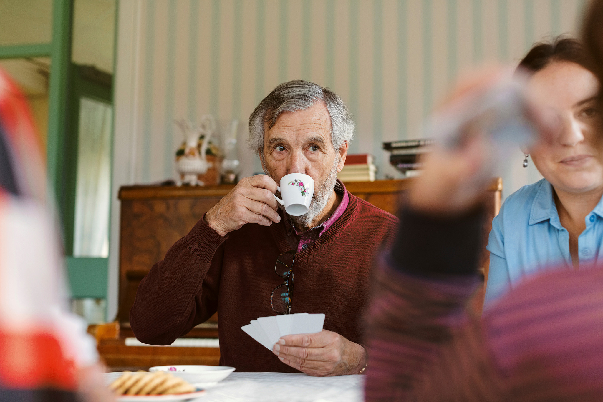Äldre man dricker kaffe ur en liten kopp och spelar kort tillsammans med några andra.
