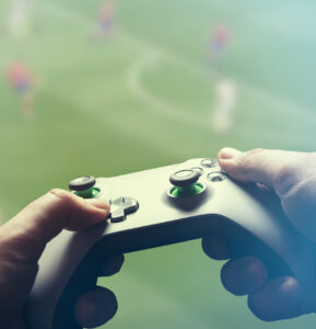 Spelkonsol i händer riktat mot en skärm med fotbollsplan och spelare i bakgrunden