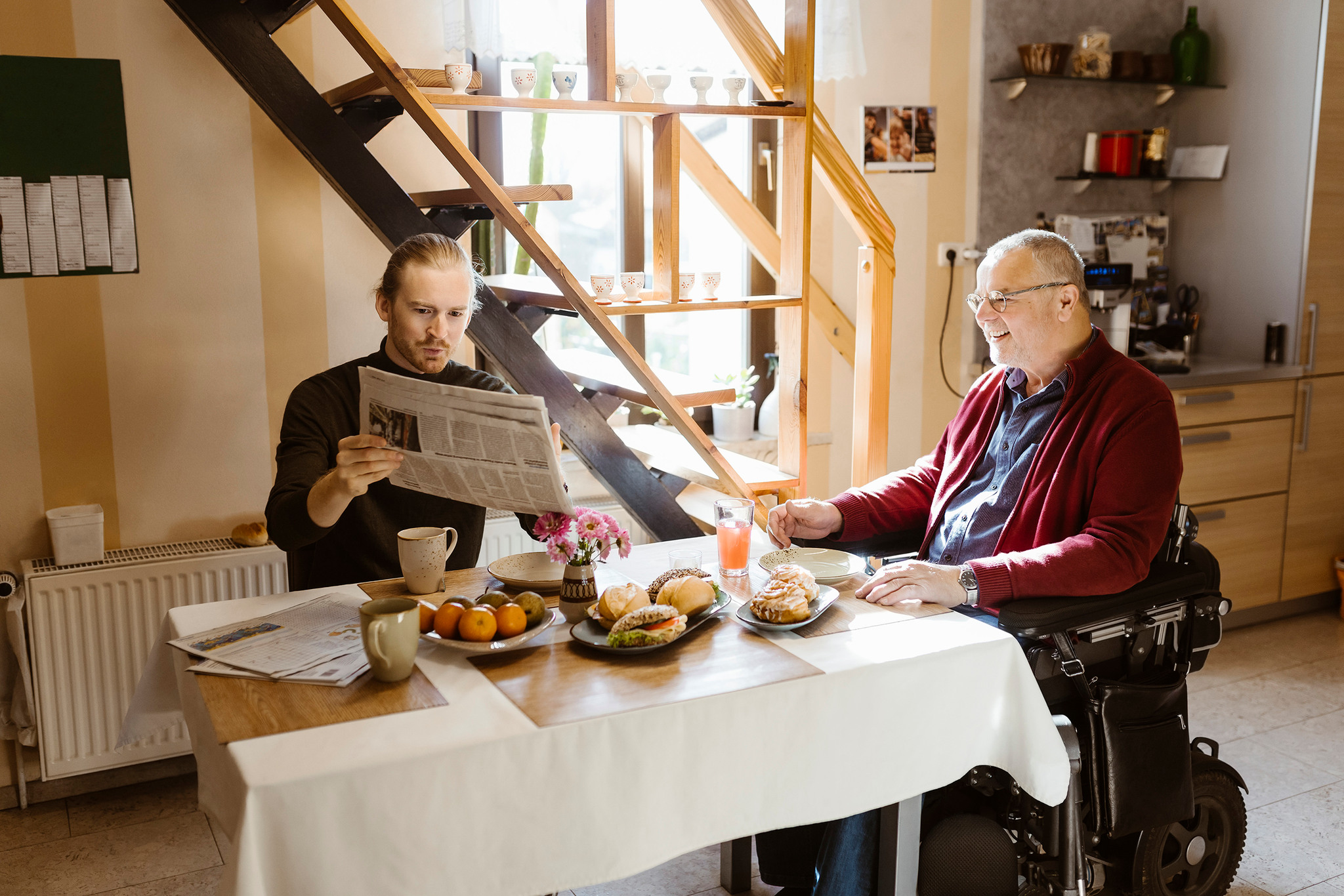 Man i rullstol vid frukostbordet tillsammans med personal som håller upp en tidning