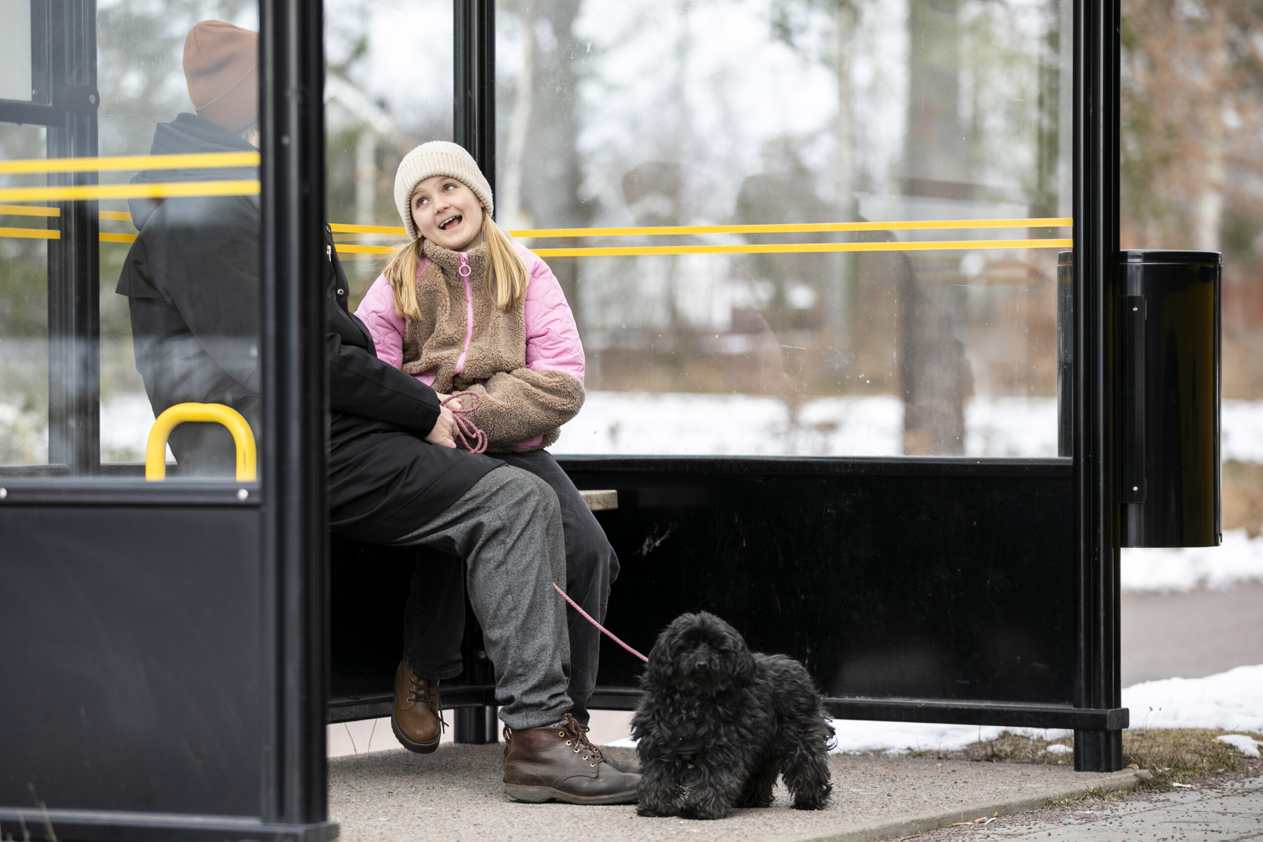 En vuxen och ett barn sitter i en glasad busskur med sin hund bredvid sig i koppel.
