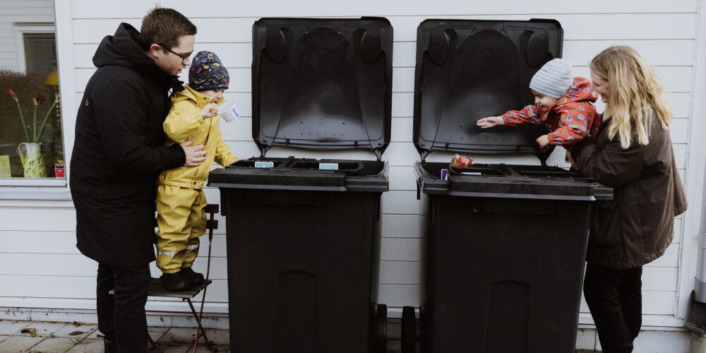 Två föräldrar slänger skräp i sopkärl tillsammans med två små barn.