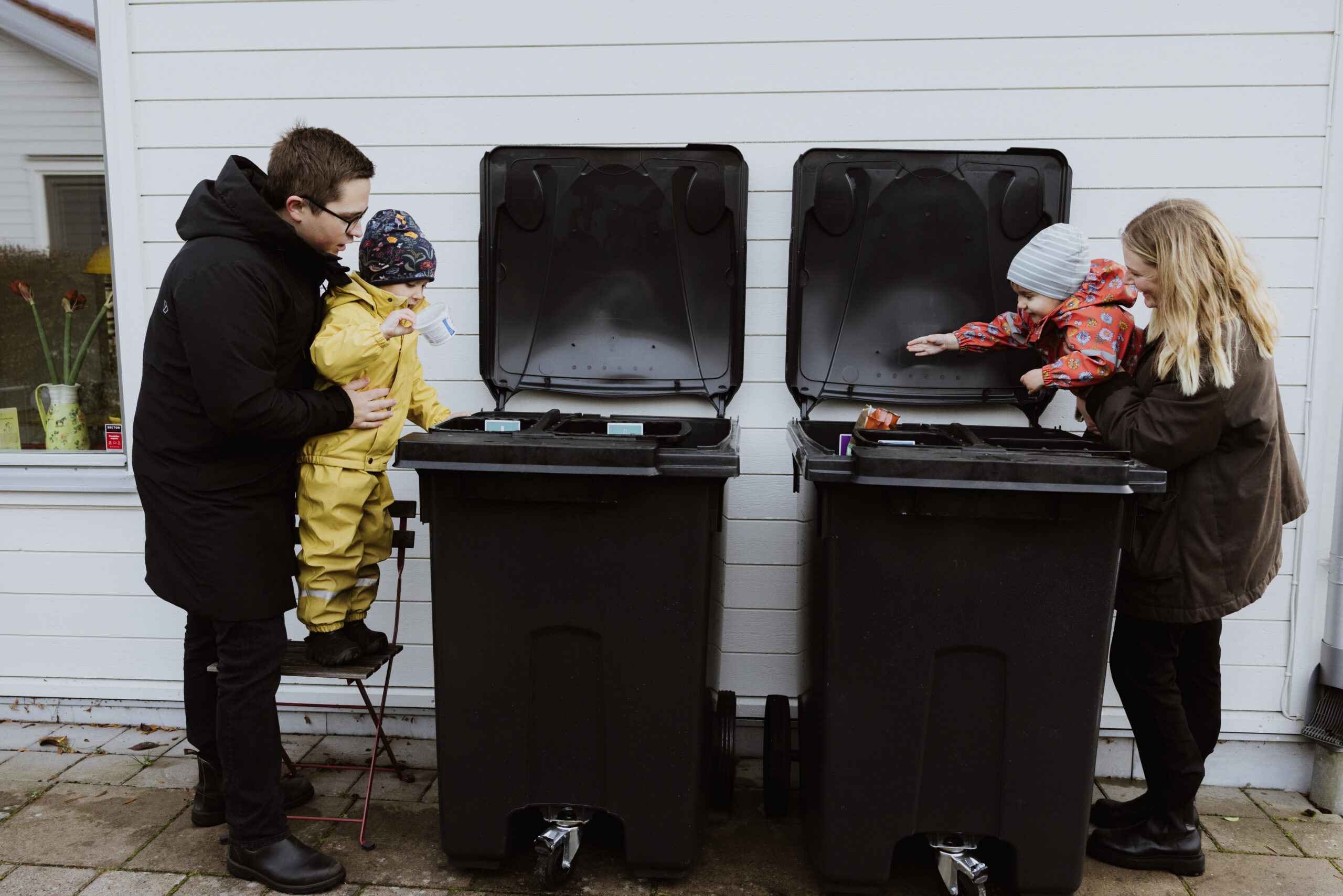 Två föräldrar slänger skräp i sopkärl tillsammans med två små barn.
