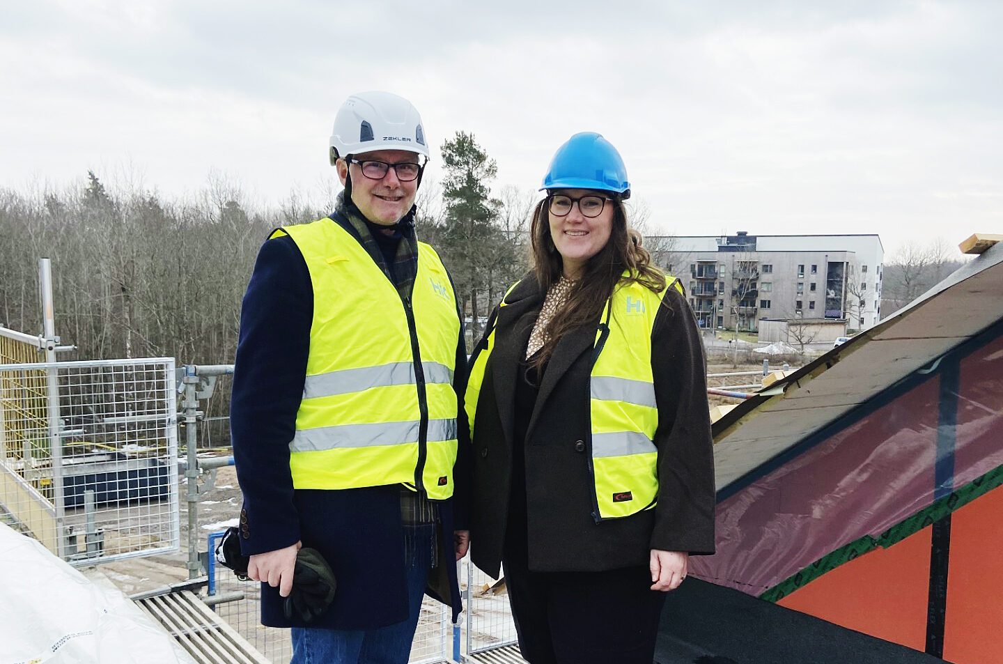 Två personer i byggarbetsplatshjälm och väst står på taket av ett nybyggt äldreboende.