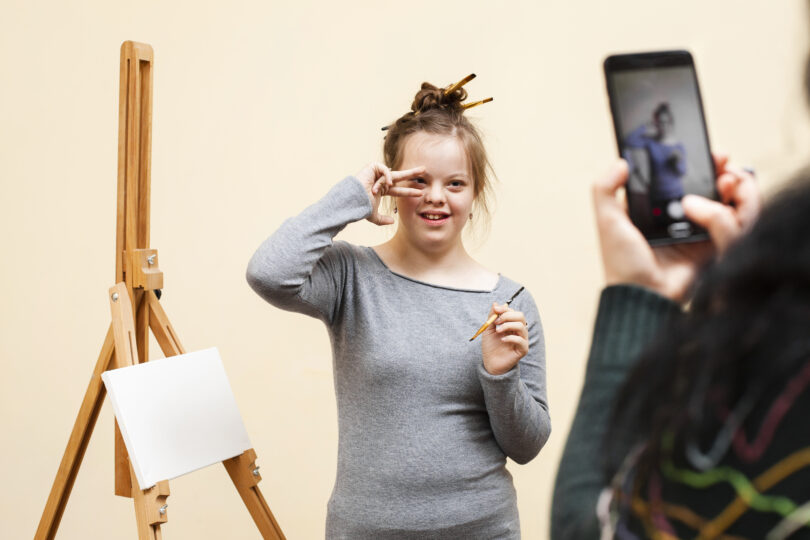 En ungdom står framför ett staffli och målar samtidigt som hon gör en pose och blir fotograferad