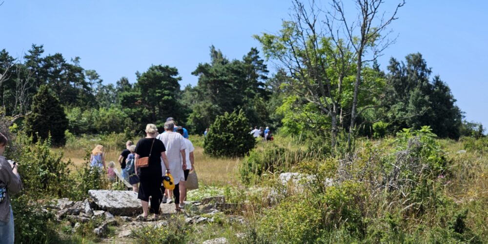 En grupp vuxna och barn vandrar bort från kameran över stenig mark genom ett öppet, buskigt landskap, med skog i bakgrunden. Solig sommardag med klarblå himmel och grönska runt stigen.
