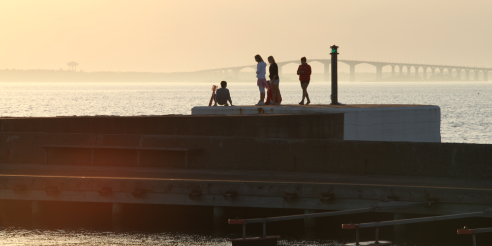 Fyra personer vistas på en vågbrytare vid havet i kvällsljus; en sitter nära en stol medan tre står och ser ut över vattnet. En grön fyrlykta syns och en lång bro skymtar i dimman.