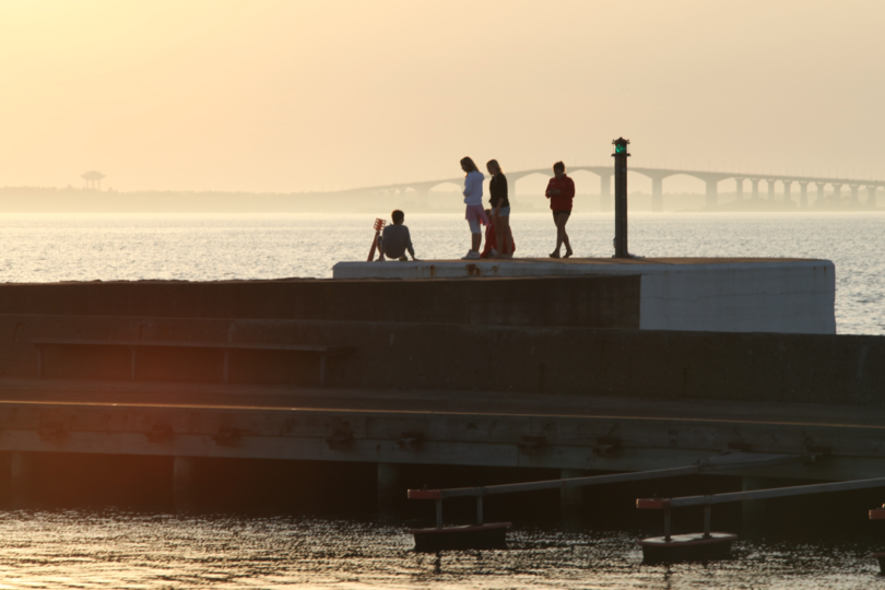 Fyra personer vistas på en vågbrytare vid havet i kvällsljus; en sitter nära en stol medan tre står och ser ut över vattnet. En grön fyrlykta syns och en lång bro skymtar i dimman.