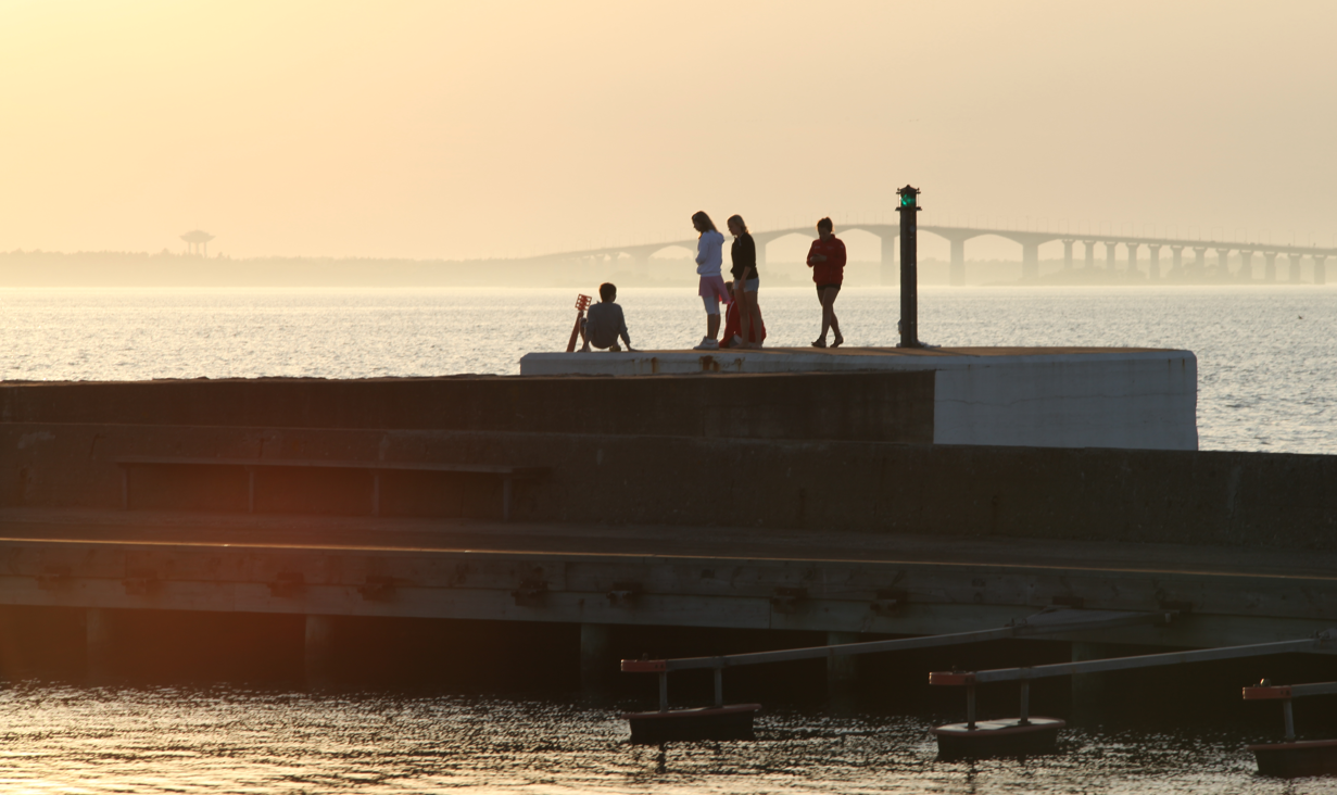 Fyra personer vistas på en vågbrytare vid havet i kvällsljus; en sitter nära en stol medan tre står och ser ut över vattnet. En grön fyrlykta syns och en lång bro skymtar i dimman.