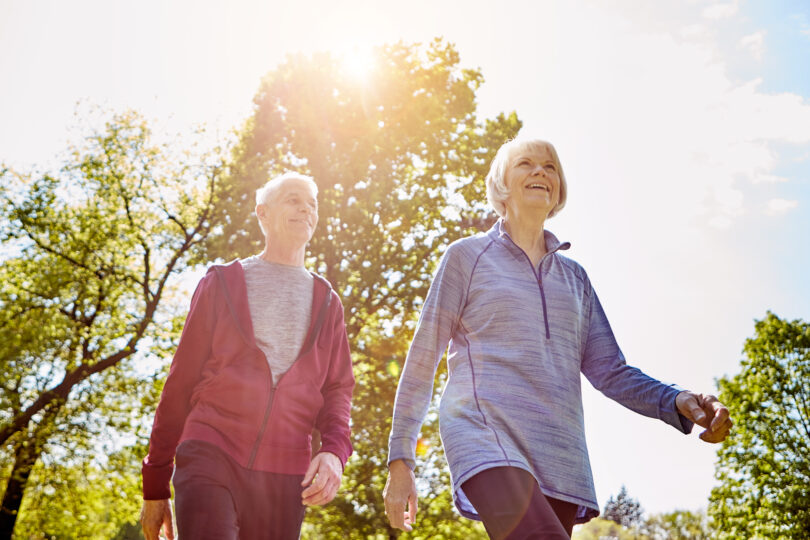 Två äldre personer promenerar i träningskläder och ser glada ut. De går sida vid sida utomhus i en grön park, med solen som lyser genom träden och skapar varmt motljus.