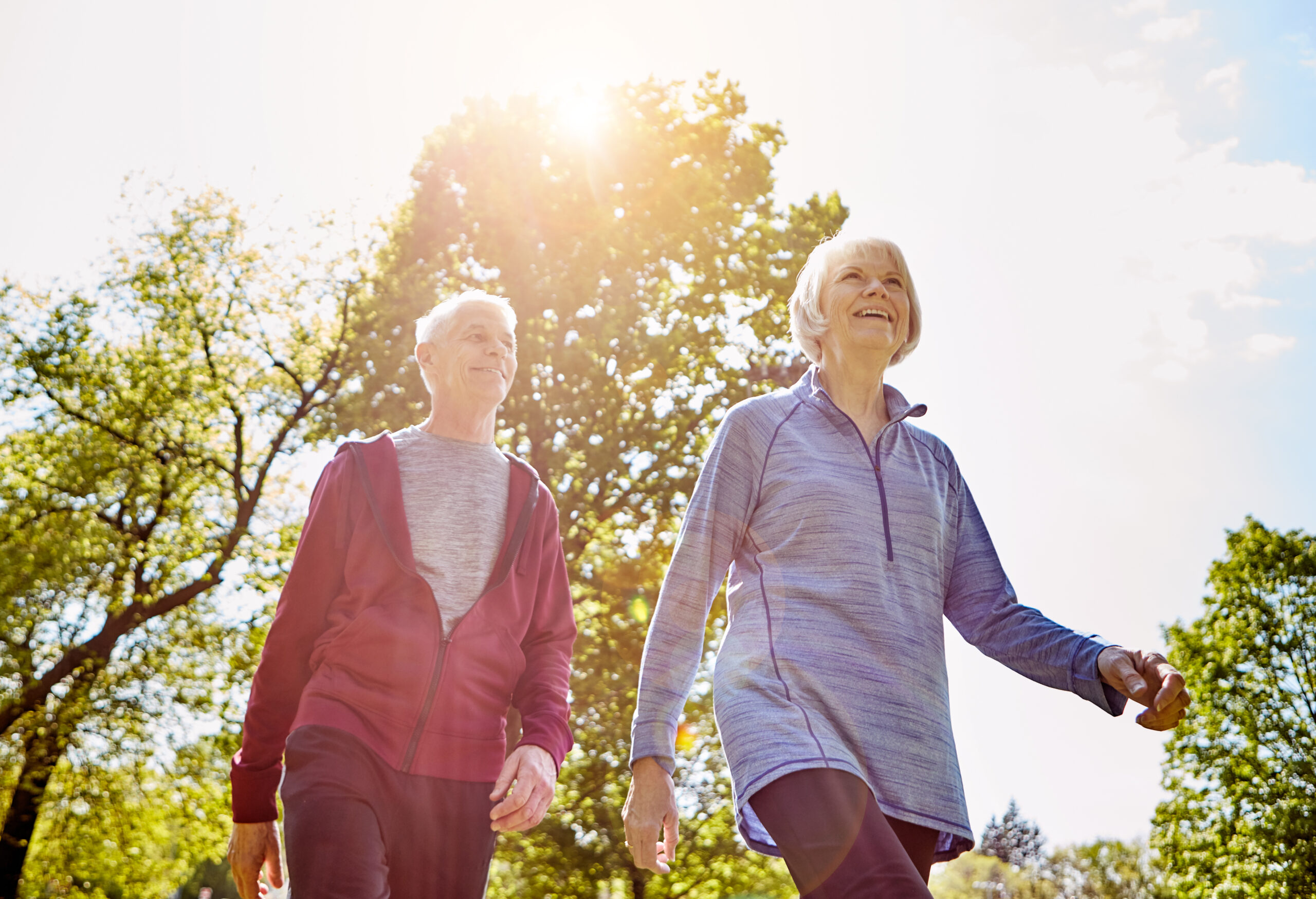 Två äldre personer promenerar i träningskläder och ser glada ut. De går sida vid sida utomhus i en grön park, med solen som lyser genom träden och skapar varmt motljus.