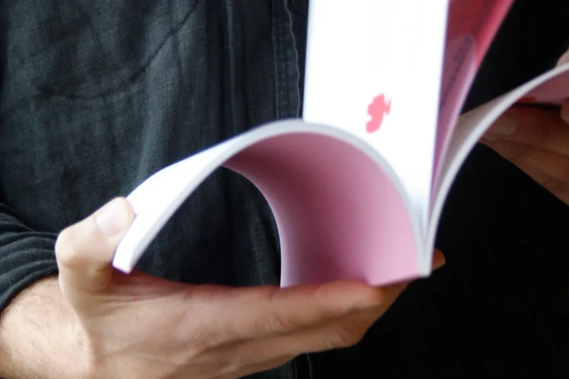 En person i mörk skjorta bläddrar i ett häfte med vita och rosa sidor, hållet öppet i händerna. Närbild som fokuserar på papperens båge och bläddringsrörelsen; ingen tydlig text syns.