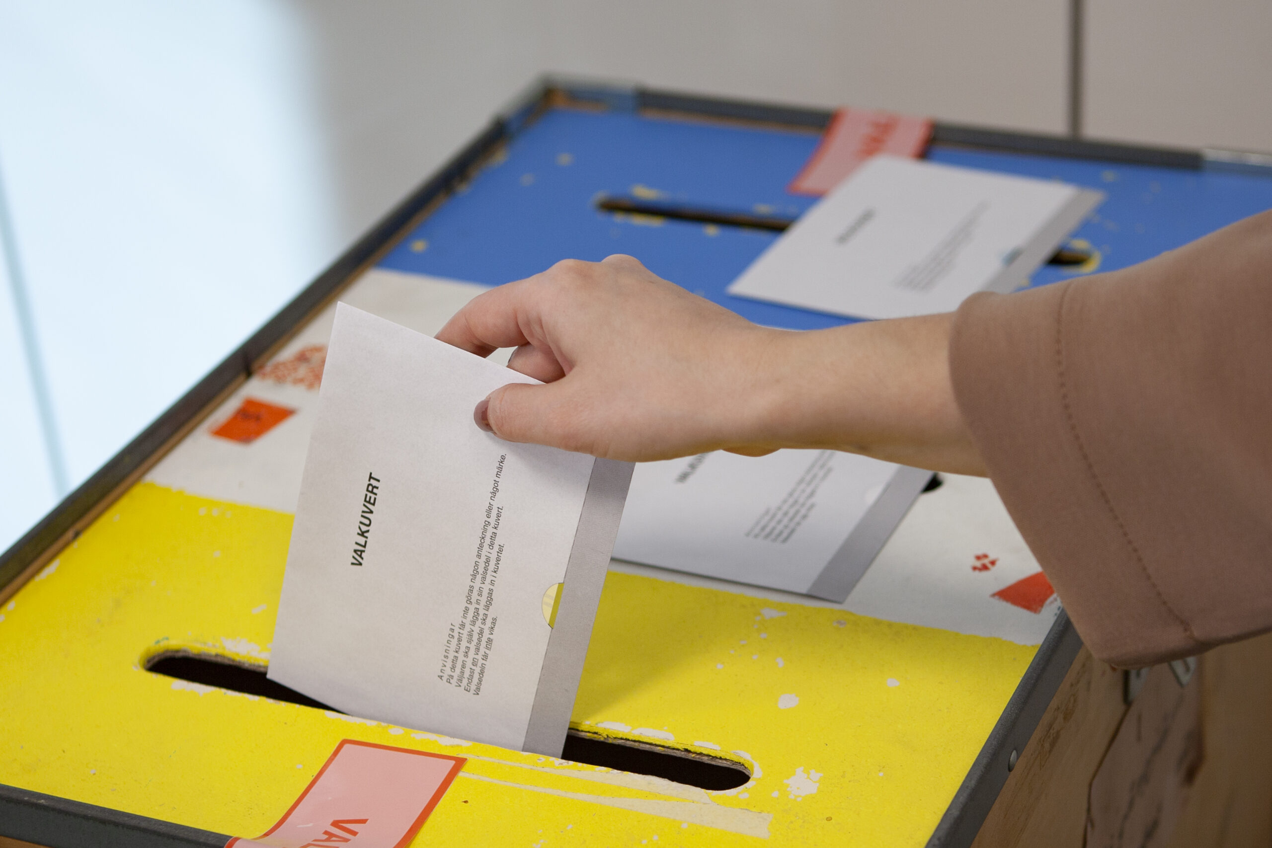 Hand stoppar ner ett kuvert i en gul och blå brevlåda med smal öppning. På kuvertet står “VALKUVERT”, och i bakgrunden syns fler papper, vilket antyder röstning eller postinlämning.