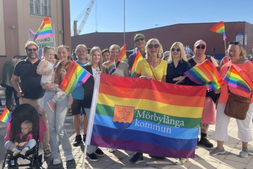Grupp människor står utomhus och poserar på en Pride-parad, håller regnbågsflaggor och en stor banderoll med texten ”Mörbylånga kommun”. Några vuxna bär barn, och en barnvagn syns i förgrunden.