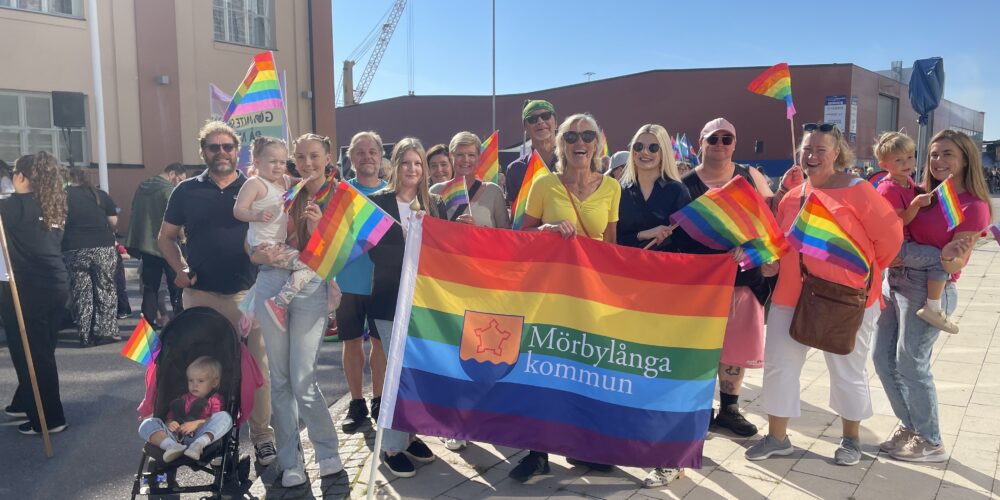 Grupp människor står utomhus och poserar på en Pride-parad, håller regnbågsflaggor och en stor banderoll med texten ”Mörbylånga kommun”. Några vuxna bär barn, och en barnvagn syns i förgrunden.