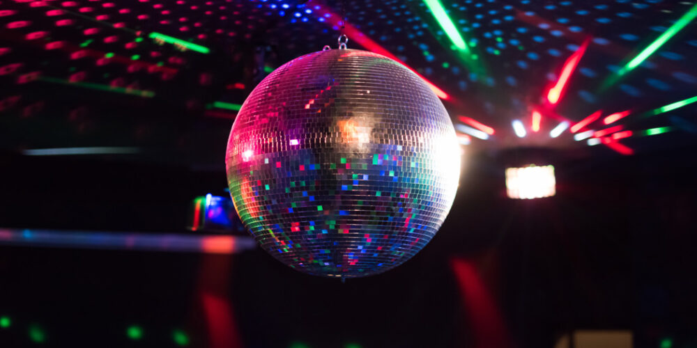 Spegelklädd discokula hänger från taket och glittrar medan den reflekterar färgade laserstrålar och ljusprickar. Mörk klubbmiljö i bakgrunden med röda, gröna och blå ljuseffekter som sprids över rummet.