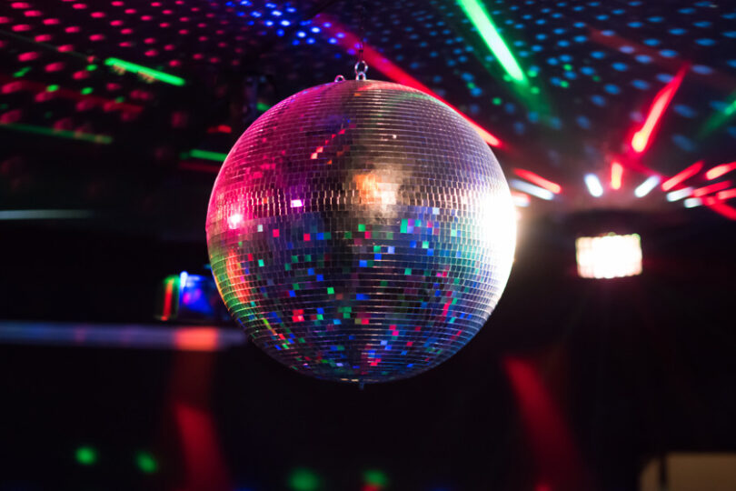 Spegelklädd discokula hänger från taket och glittrar medan den reflekterar färgade laserstrålar och ljusprickar. Mörk klubbmiljö i bakgrunden med röda, gröna och blå ljuseffekter som sprids över rummet.