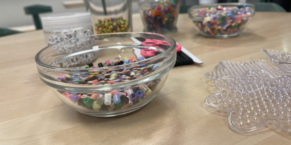 Glasskål med färgglada pärlor står på ett bord, redo att användas för pyssel. I bakgrunden syns fler burkar och skålar med pärlor samt rosa sax, och till höger en genomskinlig pärlplatta formad som ett djur.