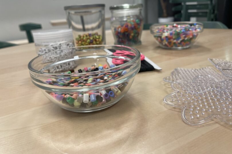 Glasskål med färgglada pärlor står på ett bord, redo att användas för pyssel. I bakgrunden syns fler burkar och skålar med pärlor samt rosa sax, och till höger en genomskinlig pärlplatta formad som ett djur.