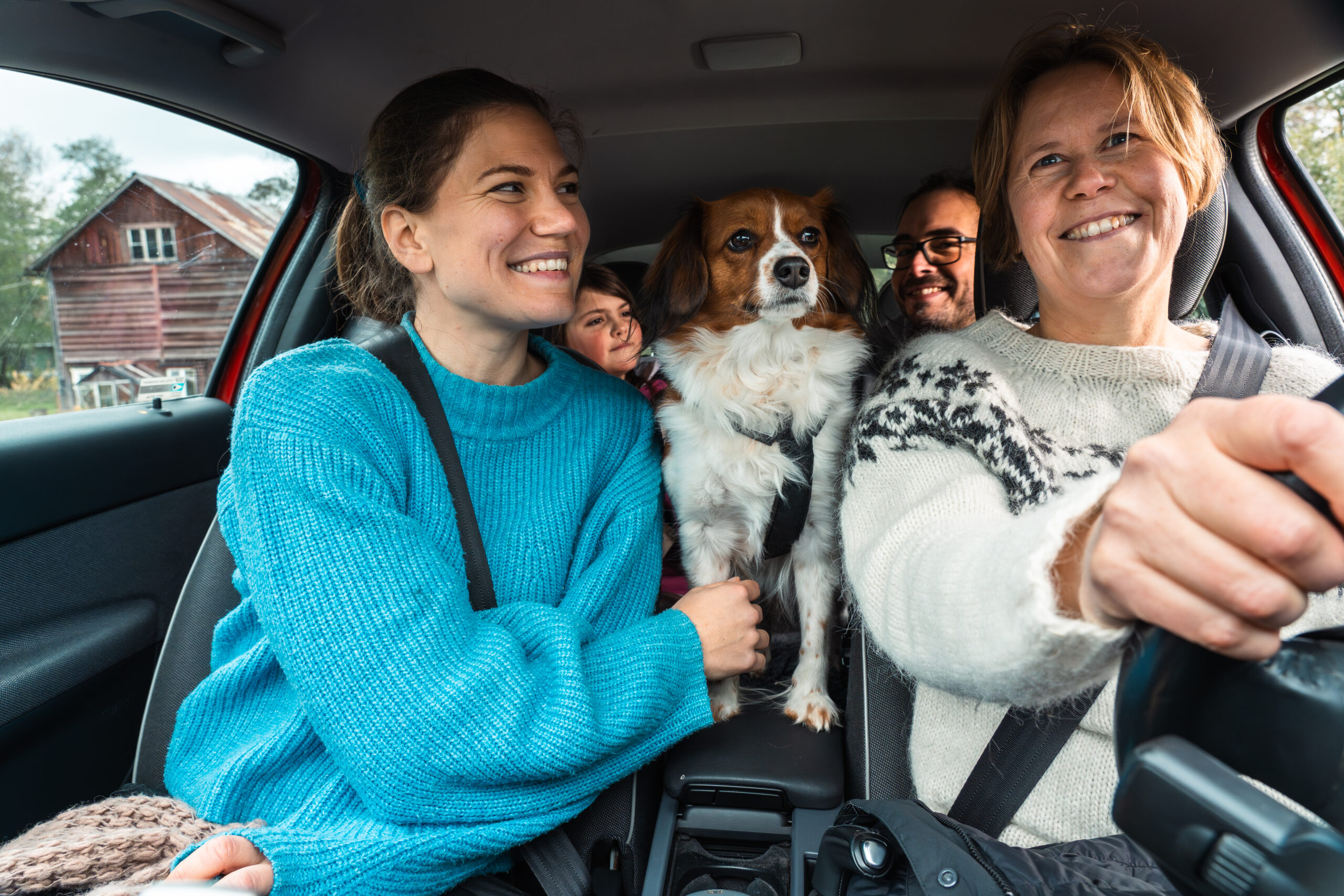 Fyra personer och en liten hund åker tillsammans i en bil; föraren och passageraren fram ler, medan hunden står mellan framsätena och tittar rakt fram. Landsbygdshus syns genom sidofönstret.
