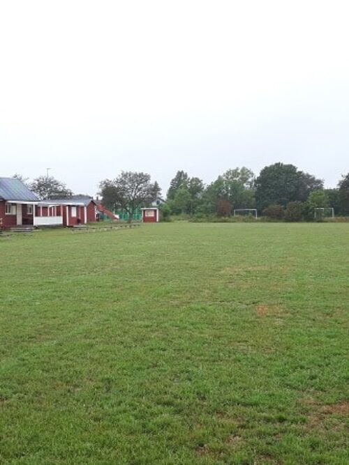 Röd klubbhusbyggnad står till vänster vid en stor gräsbevuxen fotbollsplan, med flera dörrar och skyltar på fasaden. I bakgrunden syns fotbollsmål och träd under en gråmulen himmel.