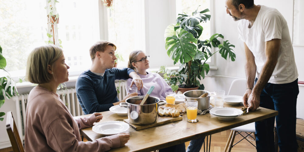 Man står vid ett dukat matbord och pratar med tre sittande personer som lyssnar. På bordet finns kastruller, tallrikar, bröd och glas med apelsinjuice, i ett ljust rum med stora fönster och gröna växter.
