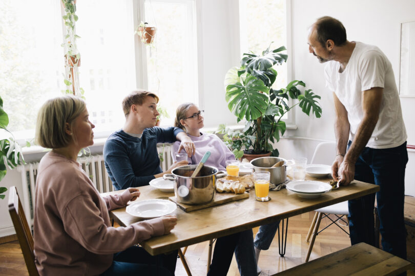 Man står vid ett dukat matbord och pratar med tre sittande personer som lyssnar. På bordet finns kastruller, tallrikar, bröd och glas med apelsinjuice, i ett ljust rum med stora fönster och gröna växter.
