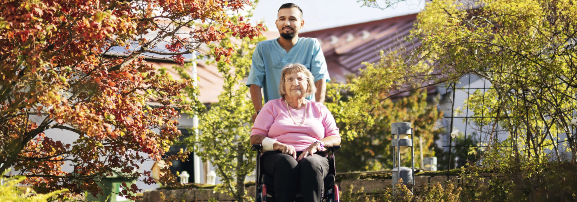 Vårdpersonal i blå arbetskläder står bakom och skjuter en äldre kvinna i rullstol längs en stenlagd gång. De är utomhus i en grönskande trädgård med träd, blå himmel och byggnader i bakgrunden.