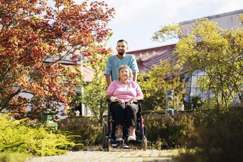 Vårdpersonal i blå arbetskläder står bakom och skjuter en äldre kvinna i rullstol längs en stenlagd gång. De är utomhus i en grönskande trädgård med träd, blå himmel och byggnader i bakgrunden.